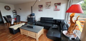 A modern living room with a black leather sofa, a wooden coffee table, and a red lamp. There is a bookshelf and a dining area in the background.