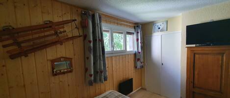 Habitació amb parets de fusta, un llit amb colcha escocesa, una finestra amb cortines amb dibuixos de cor, un armari de fusta i un televisor a sobre de l'armari.
