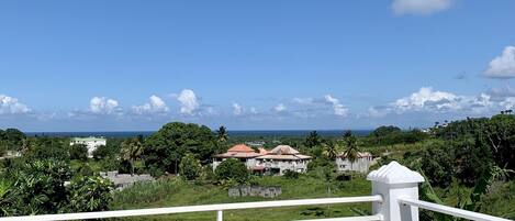 A balcony with a white railing overlooking a green landscape with houses and a clear blue sky.