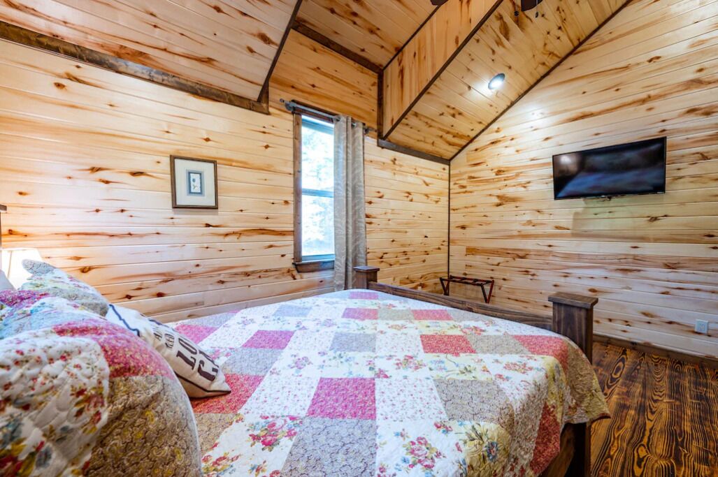 A cozy cabin-style room with wooden walls, a quilted bed, a television, and a window.