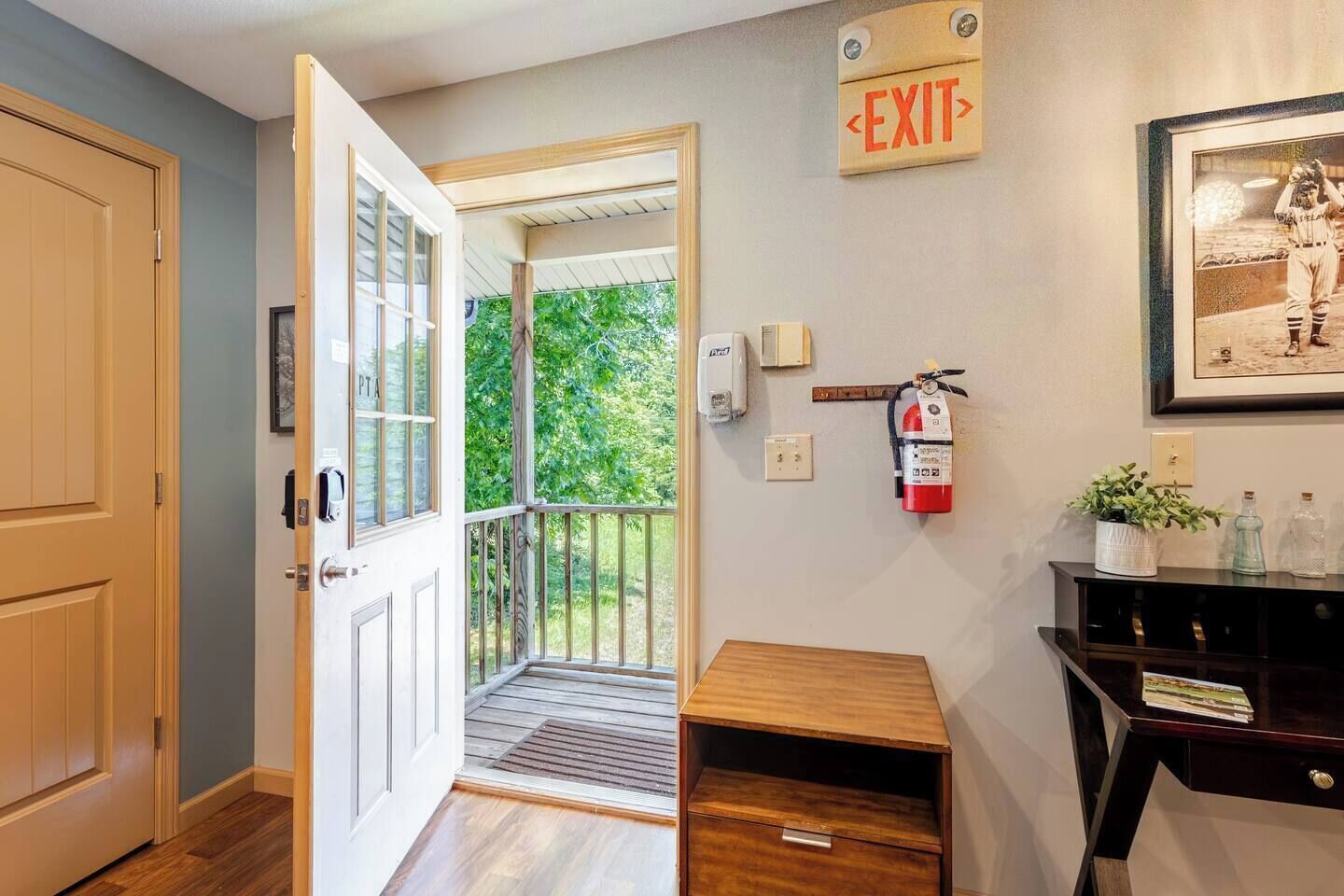 A hotel room entrance with a wooden door, an exit sign, a fire extinguisher, a small wooden table, and a framed picture on the wall.