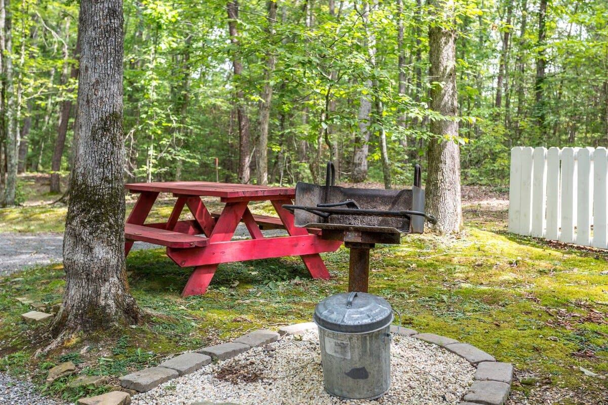 A picnic table and grill in a wooded area with a white picket fence.