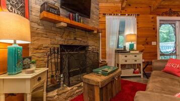 A cozy cabin interior with a stone fireplace, a television mounted on the wall, a wooden bench, and a red rug.