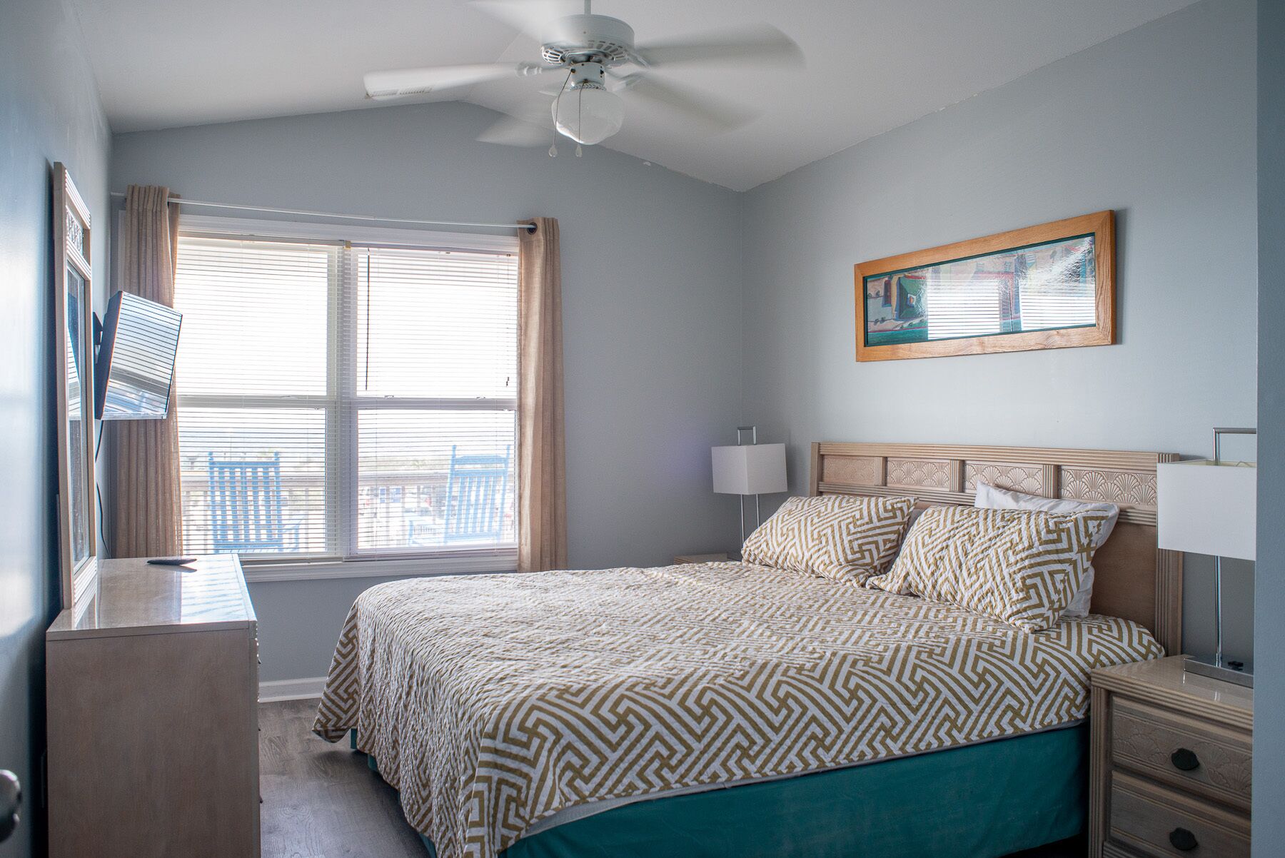 A bedroom with a bed, a nightstand, a desk, a ceiling fan, and a window with blinds.