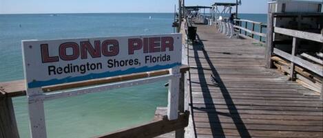 En träbrygga som sträcker sig ut i det klara, turkosfärgade vattnet, med en skylt som lyder: ”LÅNG BRYGGA, Redington Shores, Florida”.