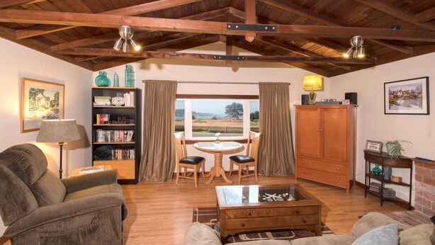 Ein gemütliches Wohnzimmer mit Holzbalkendecke, einer Bücherwand, einem Essbereich mit Tisch und Stühlen und Blick durch große Fenster.