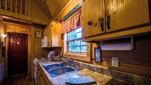 A wooden cabin-style kitchen with granite countertops, a double sink, and a window with a view of trees. - Bass Cabin with hot tub (Fort payne)