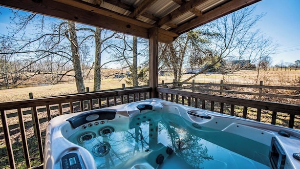 A hot tub with a control panel, surrounded by a wooden deck and metal railing, overlooking a rural landscape with trees and open fields. - Bass Cabin with hot tub (Fort payne)