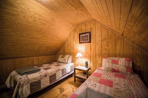 A cozy attic bedroom with two beds, wooden walls, a small table with a lamp, and a framed picture on the wall.