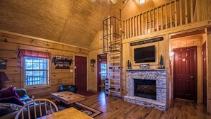 A cozy cabin interior with a stone fireplace, wooden walls, a loft area, and a television mounted above the fireplace. - Bass Cabin with hot tub (Fort payne)