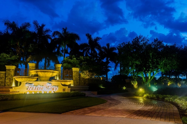 A nighttime view of a landscaped area with a fountain, palm trees, and a sign reading "Treviso Bay".