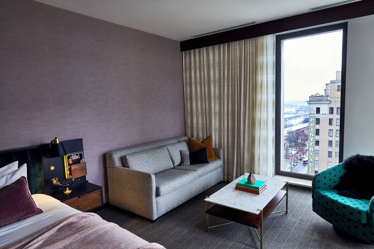 A modern hotel room with a grey sofa, a small coffee table, a teal armchair, a large window with a city view, and a minibar.