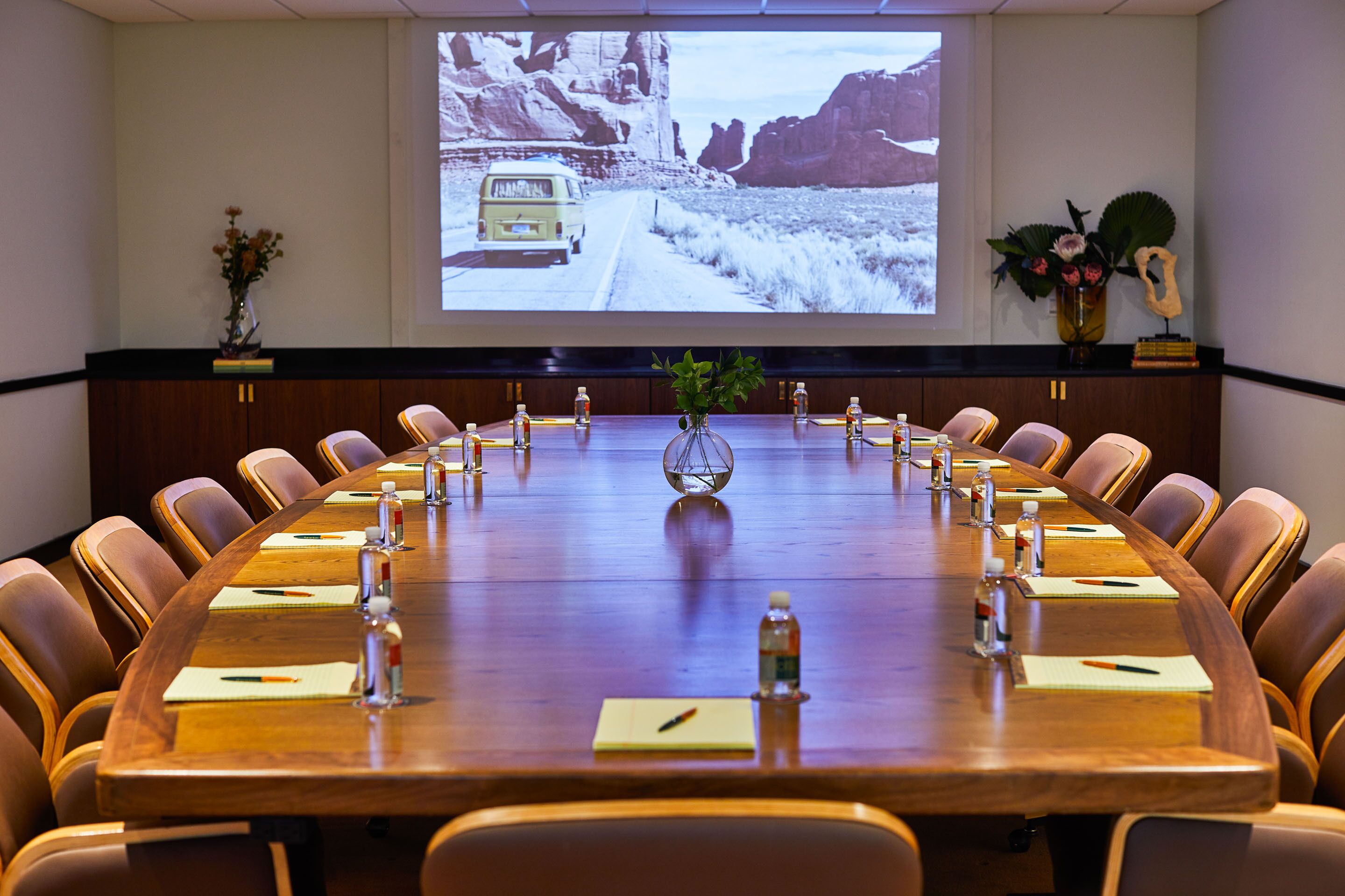 A conference room with a long table, chairs, a projector screen, and a vase with flowers.