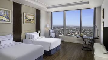 A hotel room with two beds, a desk, a chair, a lamp, and a large window offering a city view.