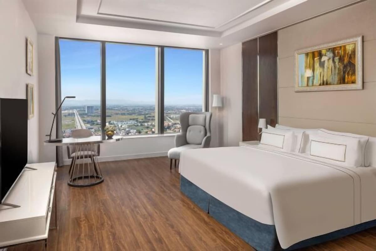 A modern hotel room with a large bed, a desk with a chair, a TV, and a large window offering a city view.