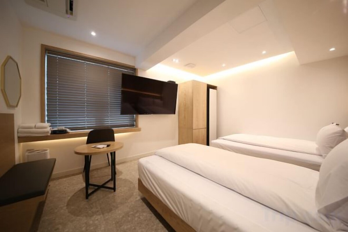 A hotel room with a bed, a small table, a chair, a TV mounted on the wall, and a window with blinds.