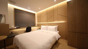 A modern hotel room with a large bed, a desk with a chair, a window with blinds, and a wooden wardrobe.