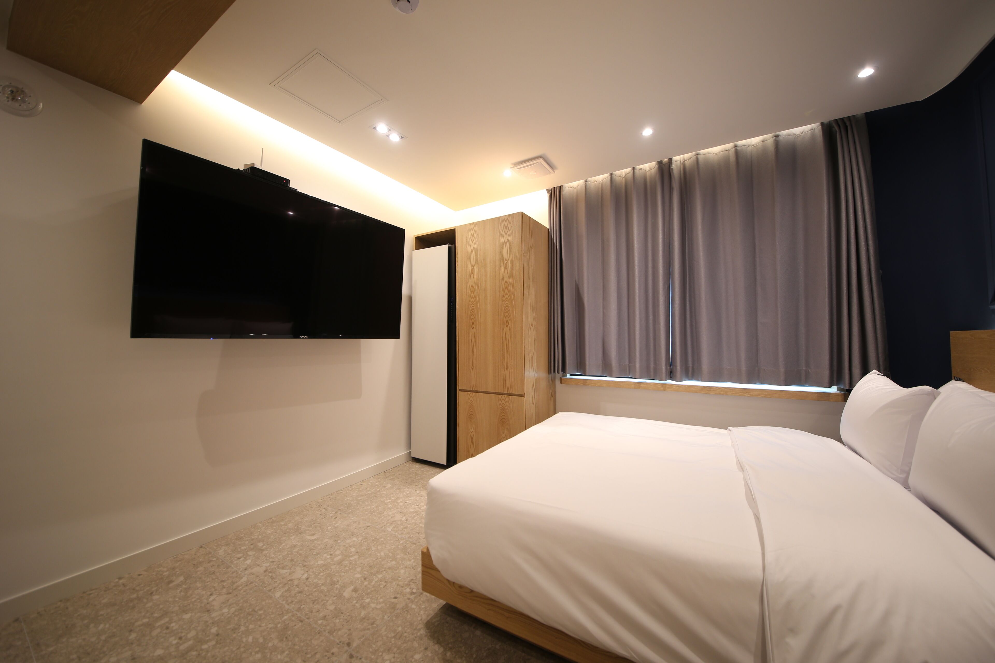 Une chambre à coucher avec un lit, un téléviseur fixé au mur, une armoire et une fenêtre avec des rideaux.