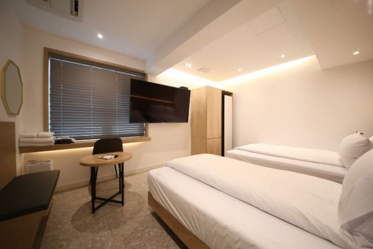Une chambre d’hôtel moderne avec deux lits, un téléviseur fixé au mur, une petite table et un banc.