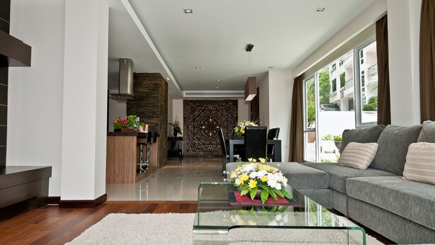 Una sala de estar moderna con un sofá gris, una mesa de centro de vidrio con flores, una cocina con salpicadero de piedra y un comedor con pared estampada.