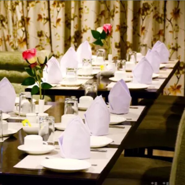 A long dining table set with white napkins, plates, and glasses, with a vase of flowers and a rose centerpiece. - Hotel Mahendra (Raipur)