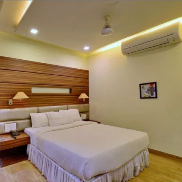 A hotel room with a wooden headboard, a large bed with white bedding, two bedside lamps, a nightstand, and an air conditioning unit on the wall. - Hotel Mahendra (Raipur)