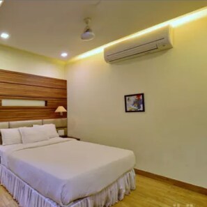 A hotel room with a large bed, wooden headboard, white bedding, a nightstand, a lamp, an air conditioning unit, and a framed picture on the wall. - Hotel Mahendra (Raipur)