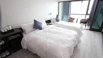 Quarto de hotel com duas camas, uma secretária pequena e uma cadeira. Possui uma janela grande com cortinas.