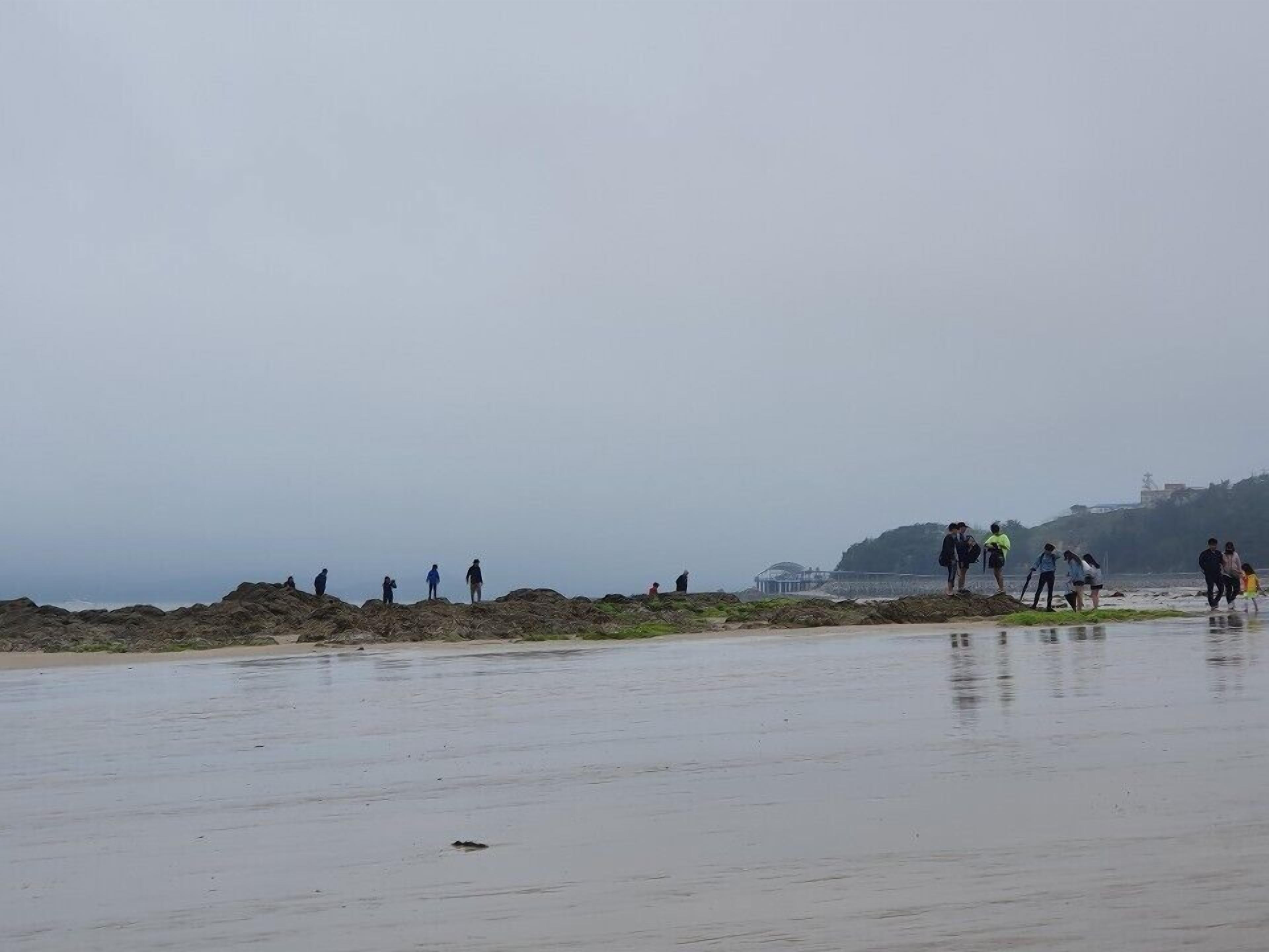 A beach with people walking on the sand and a rocky area in the background.