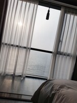 A room with a bed, a window with sheer curtains, and a view of the sea.