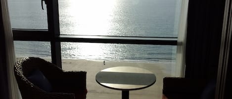 A room with a large window overlooking a beach, a small round table, and two wicker chairs.