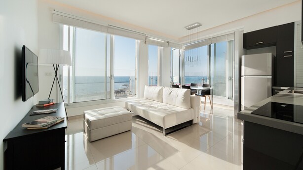 A modern living room with a white sofa, a black side table, and a large window overlooking the sea. - Sunset Panorama Apartment by Sea N' Rent (Tel Aviv)