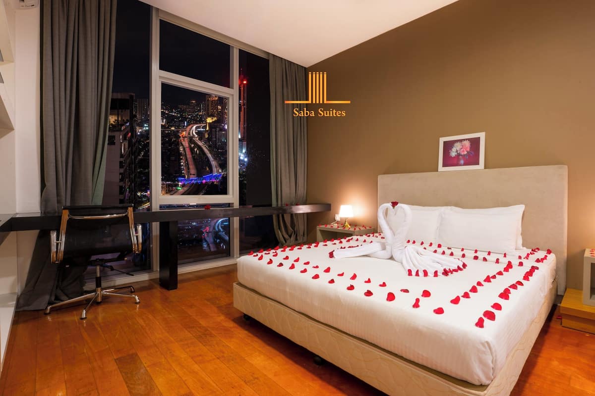 A hotel room with a bed adorned with rose petals and a swan-shaped towel, a desk with a chair, a nightstand with a lamp, and a view of a cityscape at night.
