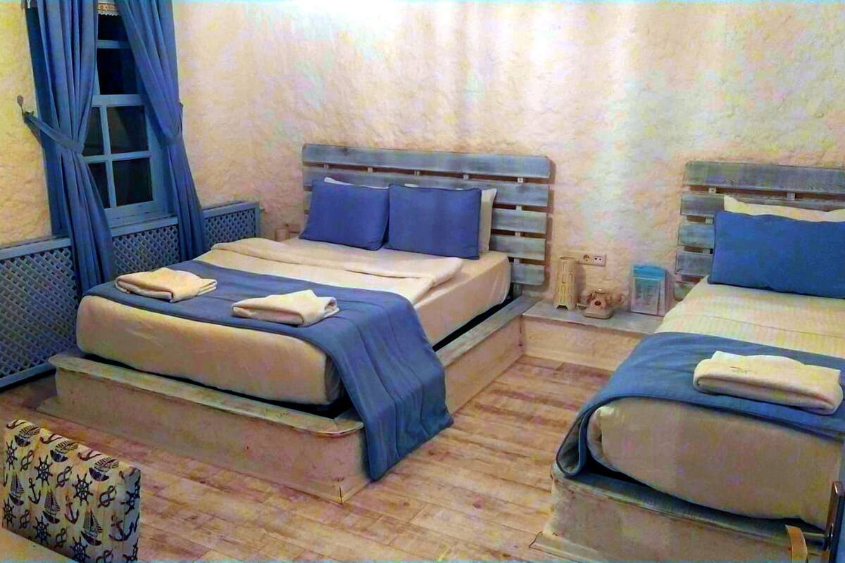 A room with two beds, each with blue bedding and pillows, a small table with a lamp, and a window with curtains.