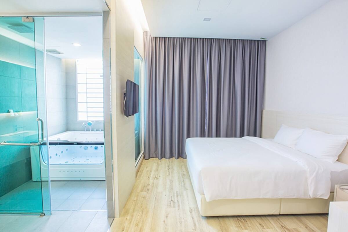 A modern hotel room with a large bed, a glass-enclosed shower area, and a view of the city.