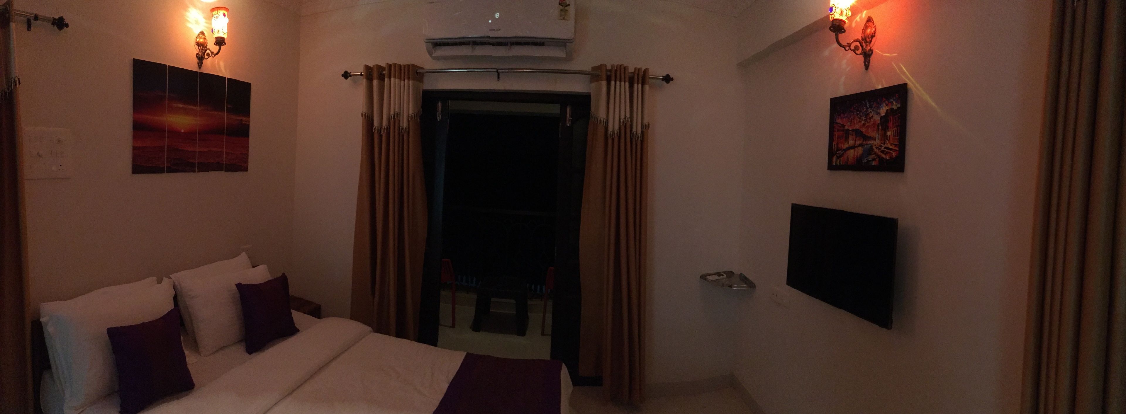 Une chambre d’hôtel comprenant un lit, deux climatiseurs fixés au mur, une porte avec des rideaux et deux cadres d’images accrochés au mur.