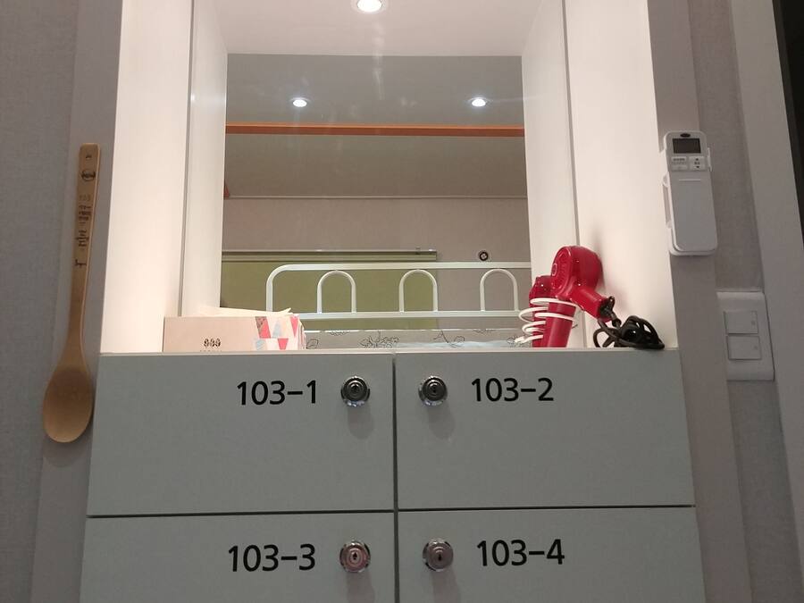 Une salle de bain avec un miroir, quatre tiroirs numérotés de 103-1 à 103-4, un sèche-cheveux rouge et une cuillère en bois accrochée au mur.