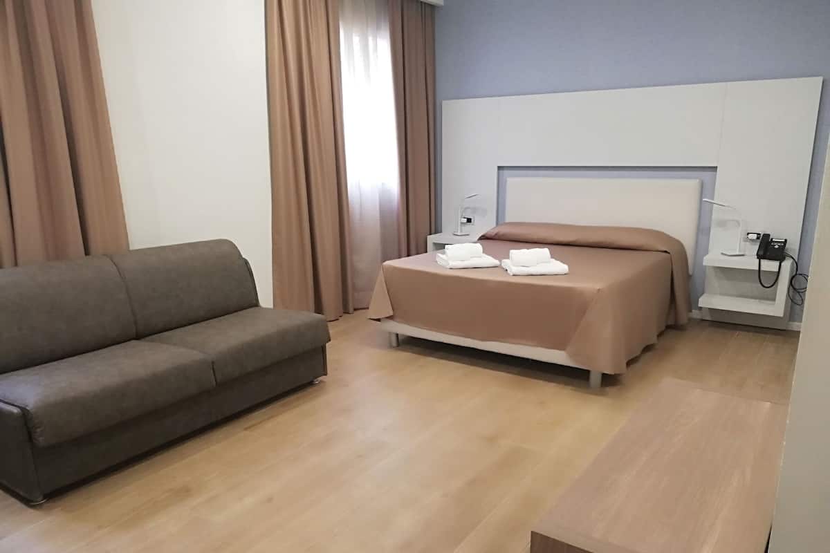 A modern hotel room with a sofa, a bed with a headboard, a bedside table with a lamp, and a phone on a shelf.