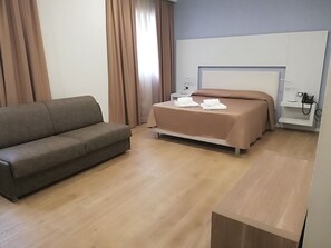 A modern hotel room with a sofa, a bed with a headboard, a bedside table with a lamp, and a phone on a shelf.