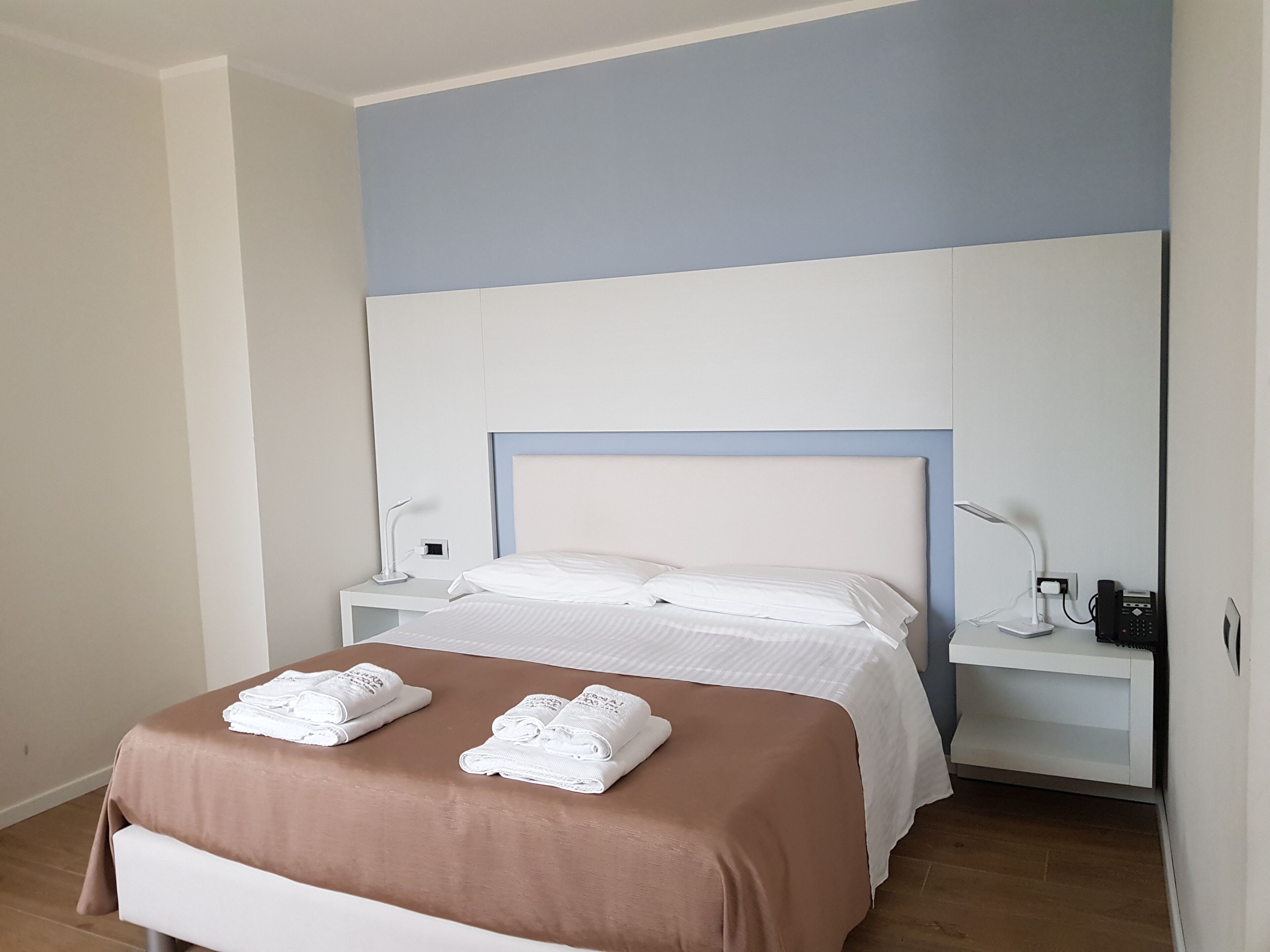 A neatly made bed with white linens and two folded towels, a bedside table with a lamp, and a nightstand with a phone and charger.