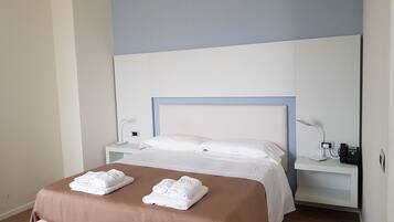 A neatly made bed with white linens and two folded towels, a bedside table with a lamp, and a nightstand with a phone and charger.