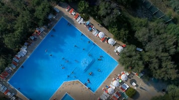 An aerial view of a rectangular swimming pool surrounded by beach umbrellas and lounge chairs, with a forested area in the background.