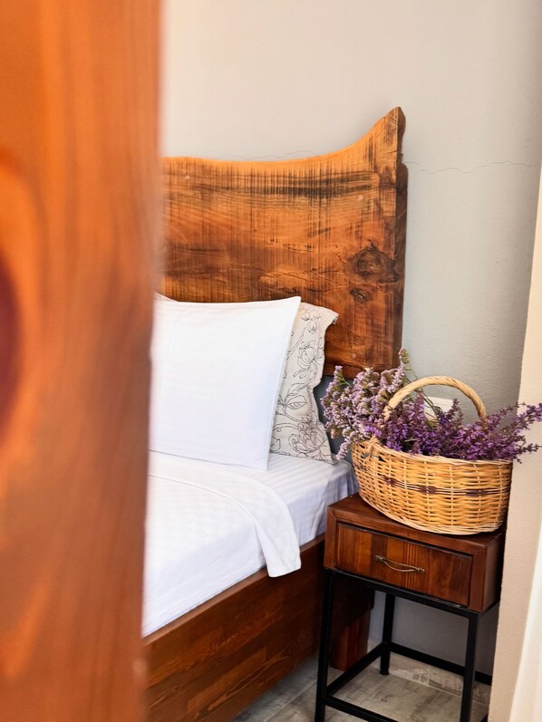 A bed with a wooden headboard, a white pillow, a patterned pillow, a white blanket, and a wicker basket with purple flowers on a bedside table. - Tenedion Hotel (Bozcaada)