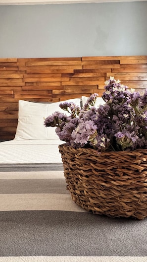 Una cama con cabecera de madera, una cesta tejida con flores moradas y una colcha a rayas.