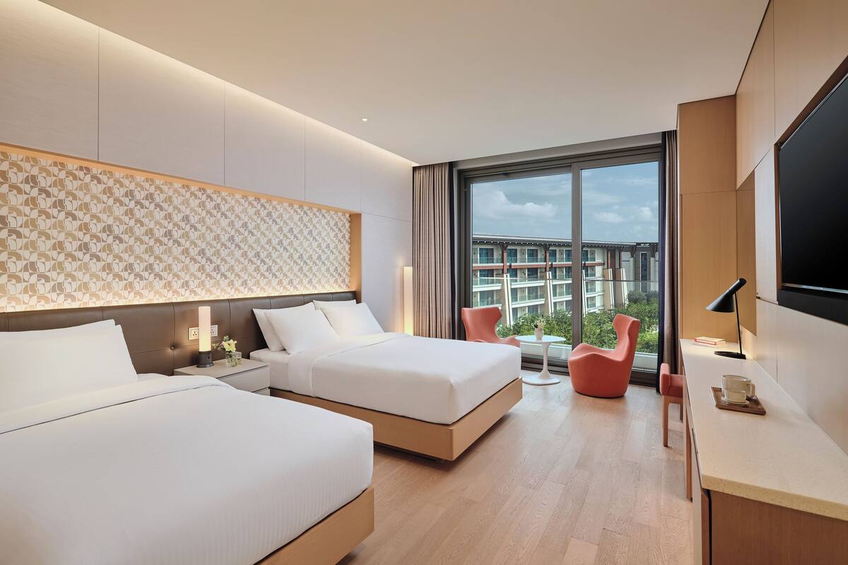 A modern hotel room with two beds, a desk, a chair, and a large window offering a city view.