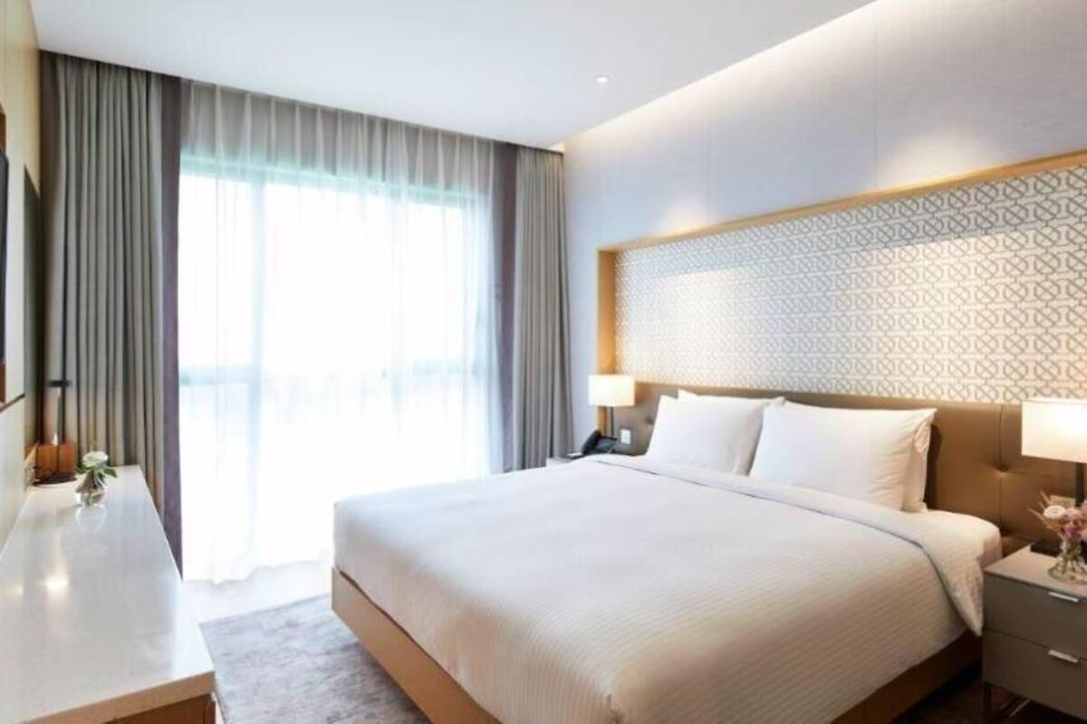 A modern hotel room with a large bed, bedside tables, a desk, and a large window with curtains.
