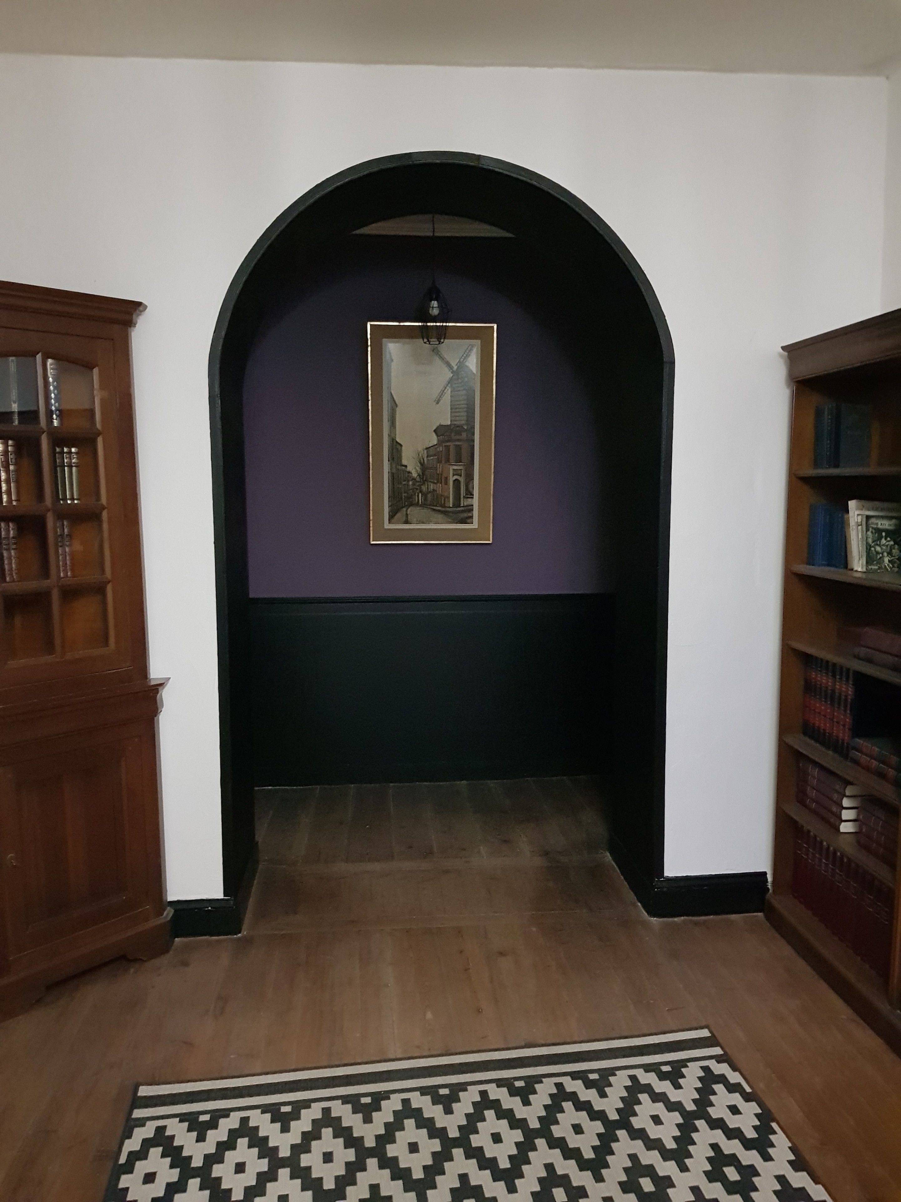 A hallway with a wooden floor, a patterned rug, a framed painting on the wall, and wooden bookshelves on either side.