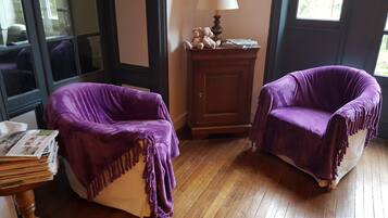 Twee fauteuils met paarse bekleding, een houten kast en een lamp op een tafel met tijdschriften.