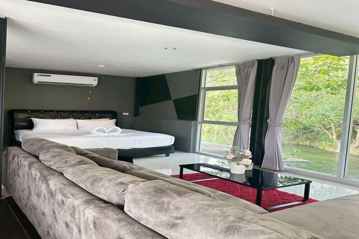 A modern bedroom with a large bed, a sofa, and a view of greenery through large windows.
