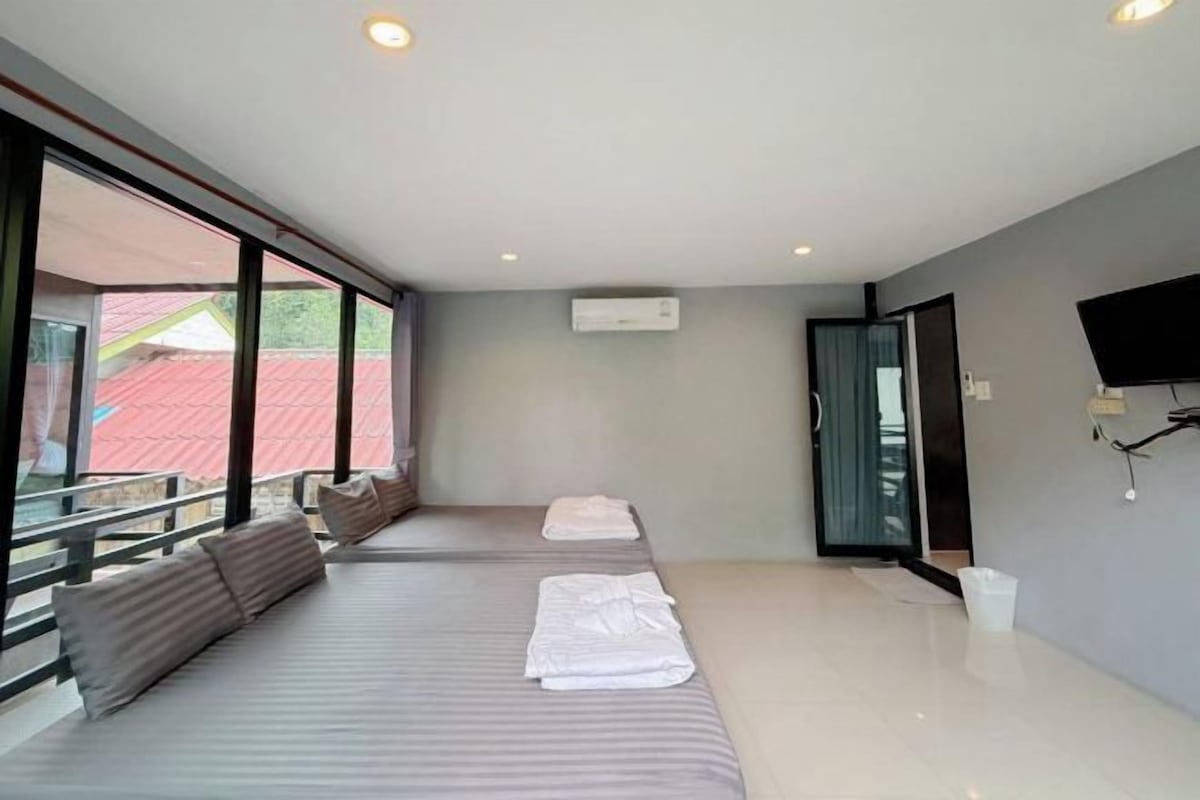 A modern hotel room with a large window, a flat-screen TV mounted on the wall, and a balcony with a red roof.